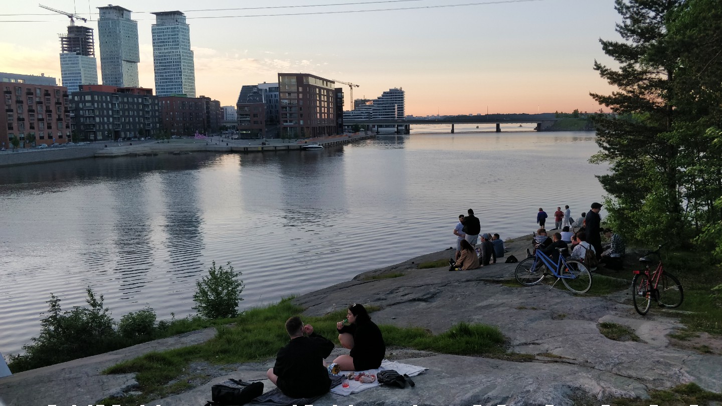 People are sitting on the rock at Mustikkamaa seaside. Apartment buildigns of Kalasatama rise on the other side of the shore.