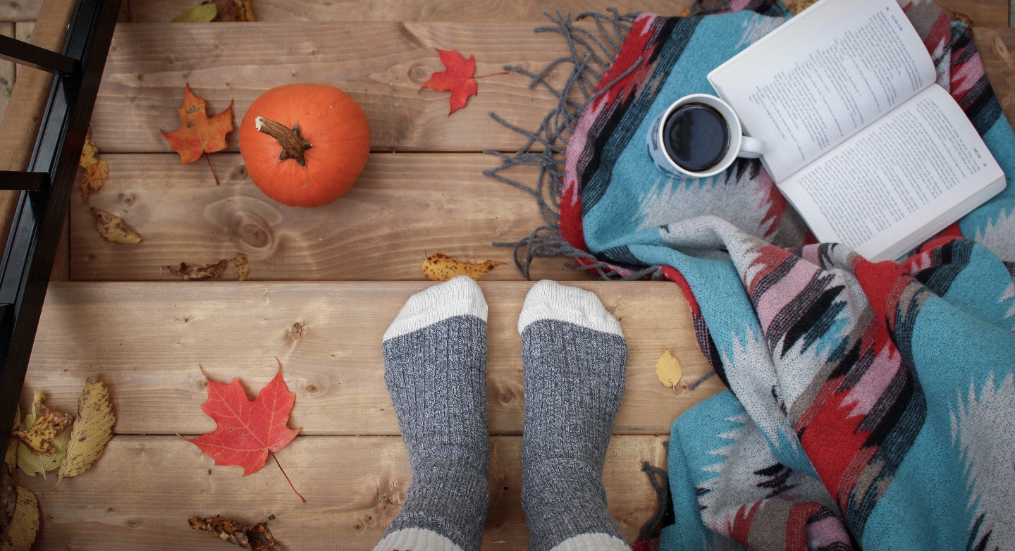 On a wooden stairs lay autums leafs, a tiny pumpkin, a woollen blanket, a coffee mug and an open book. Some one stands on the stairs. Only the wollen socks can be seen.