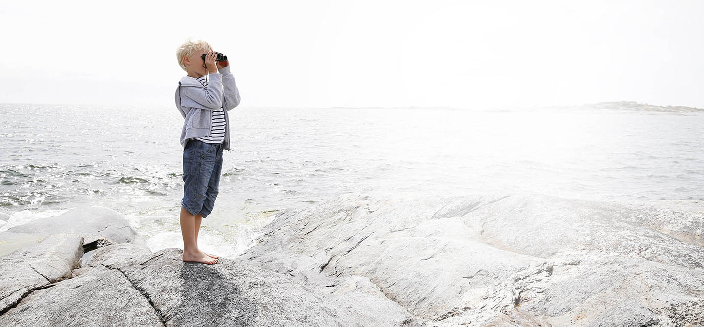 En pojke står på en sten vid havet och tittar i en kikare.